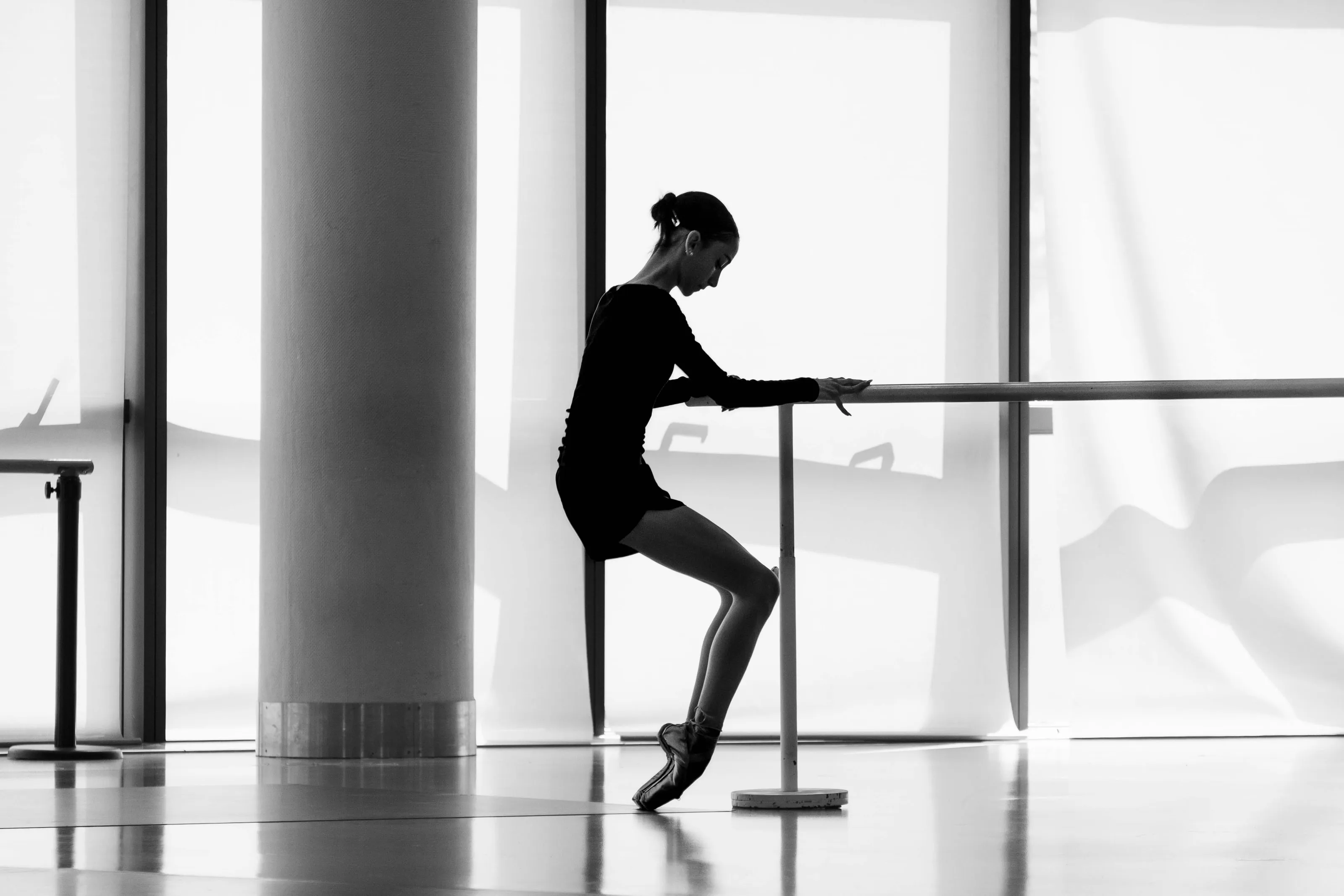 A ballerina in silhouette at the barre, en pointe, before tall windows