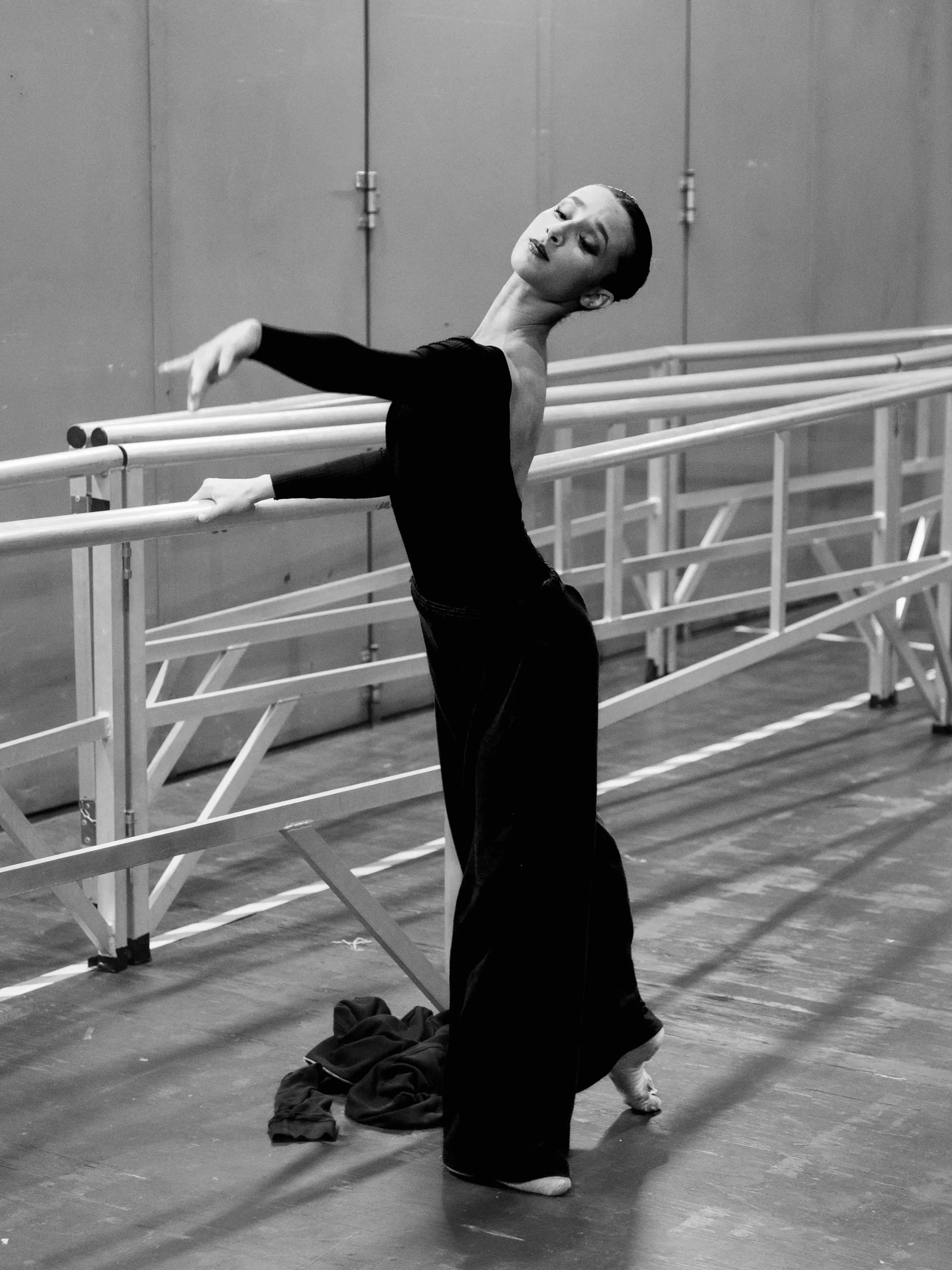 A ballerina in black stretches at the barre backstage