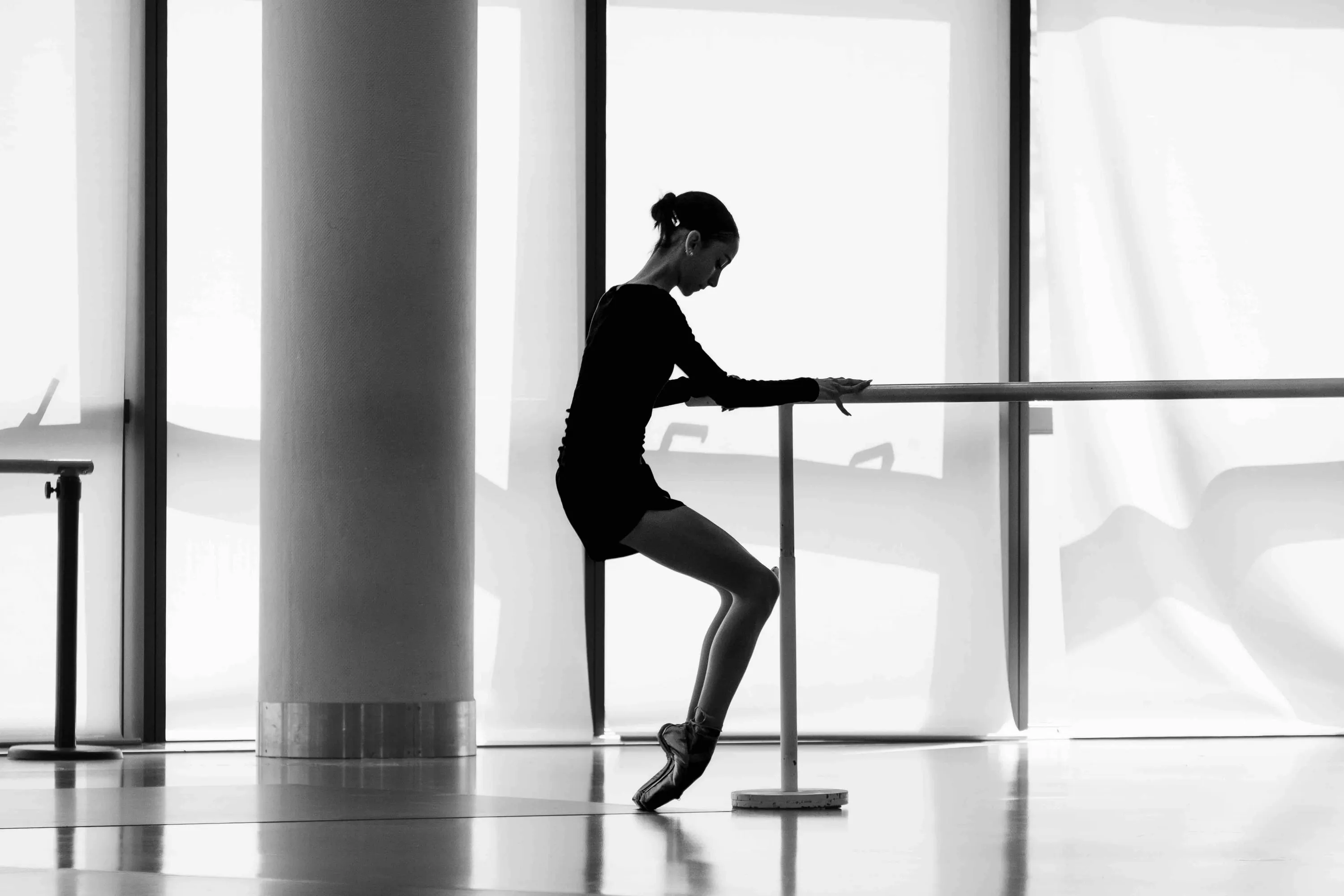 A ballerina in silhouette at the barre, en pointe, before tall windows