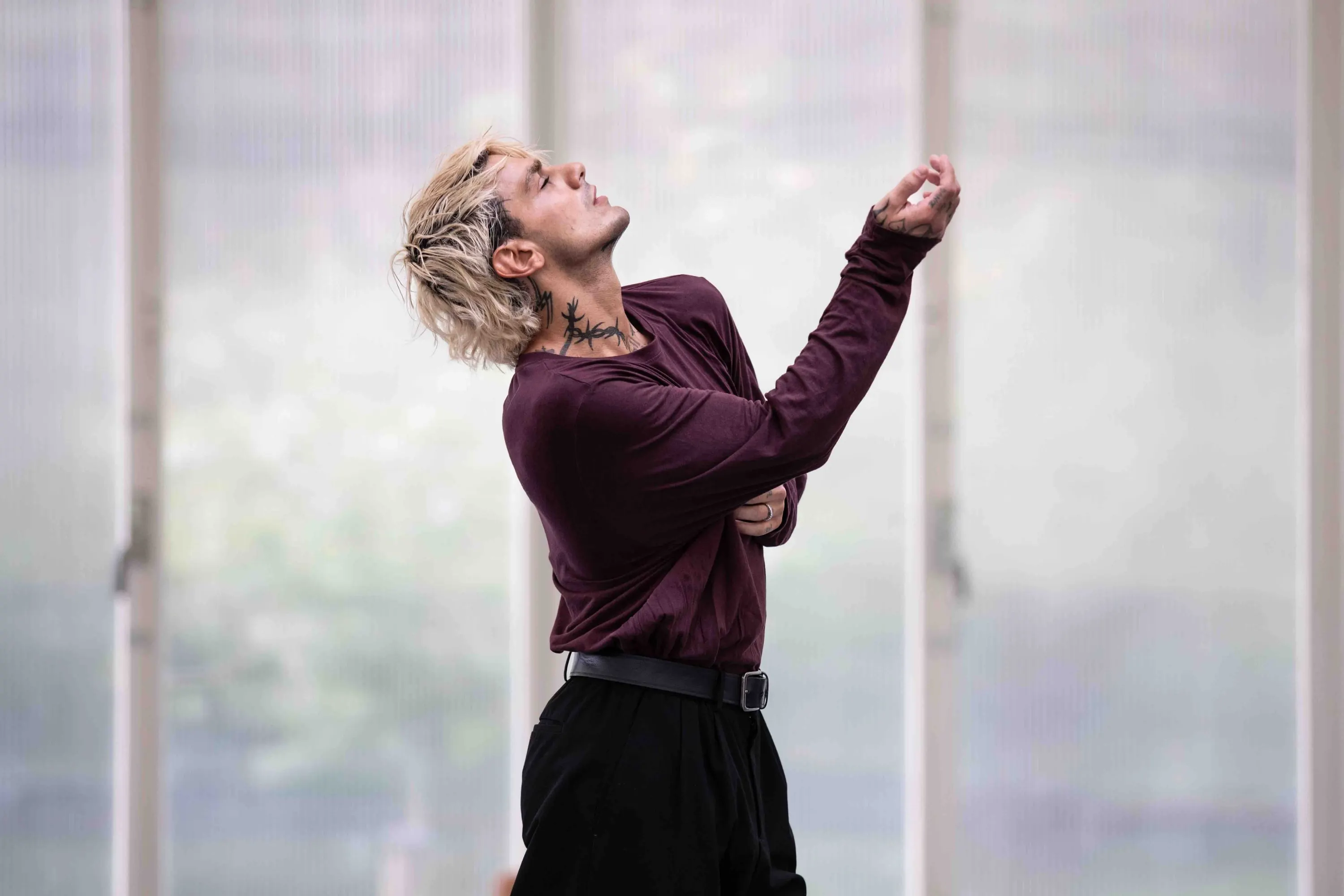 A dancer in dark red raises an arm before the window