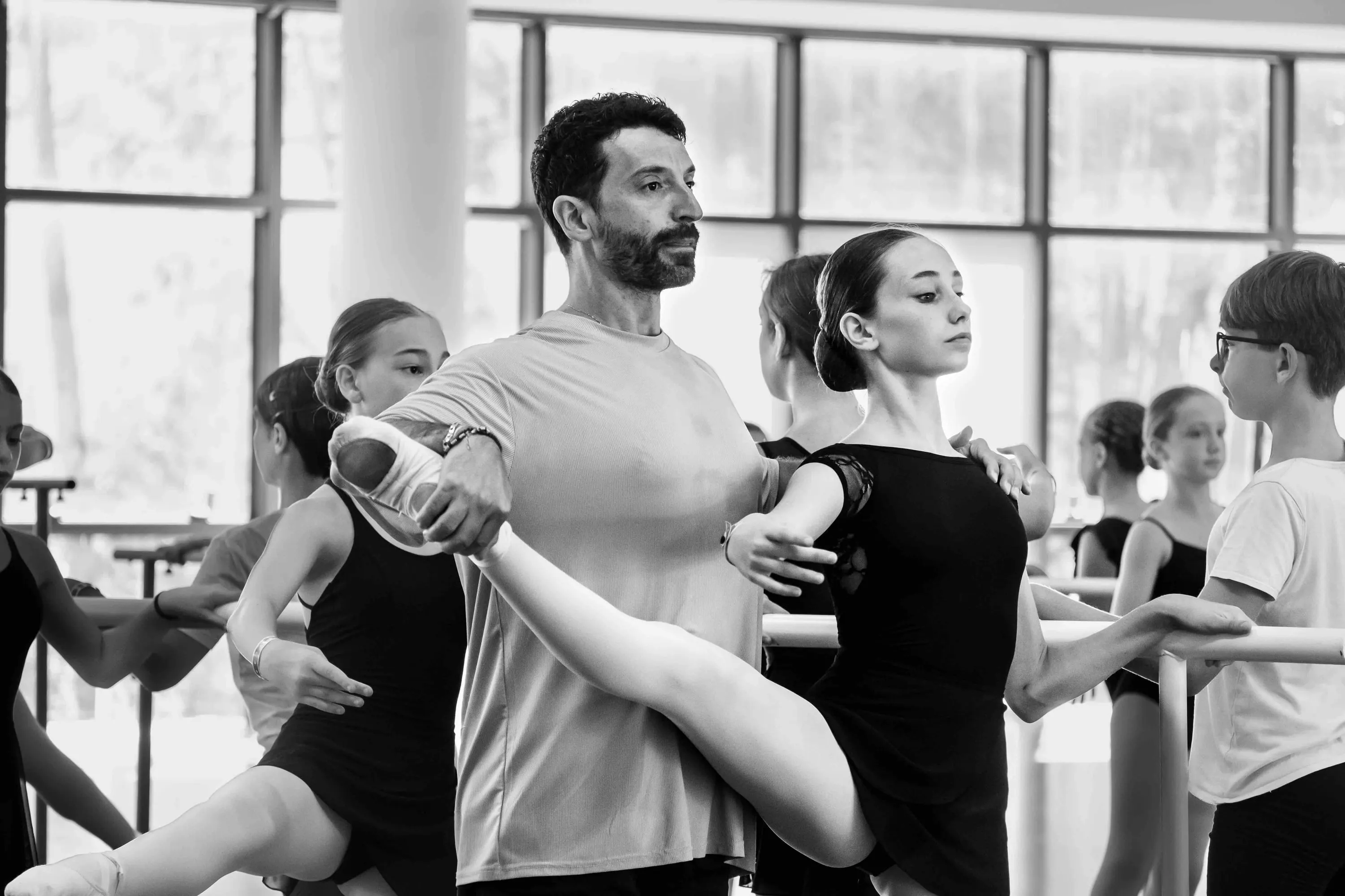The teacher adjusts a student's leg at the barre