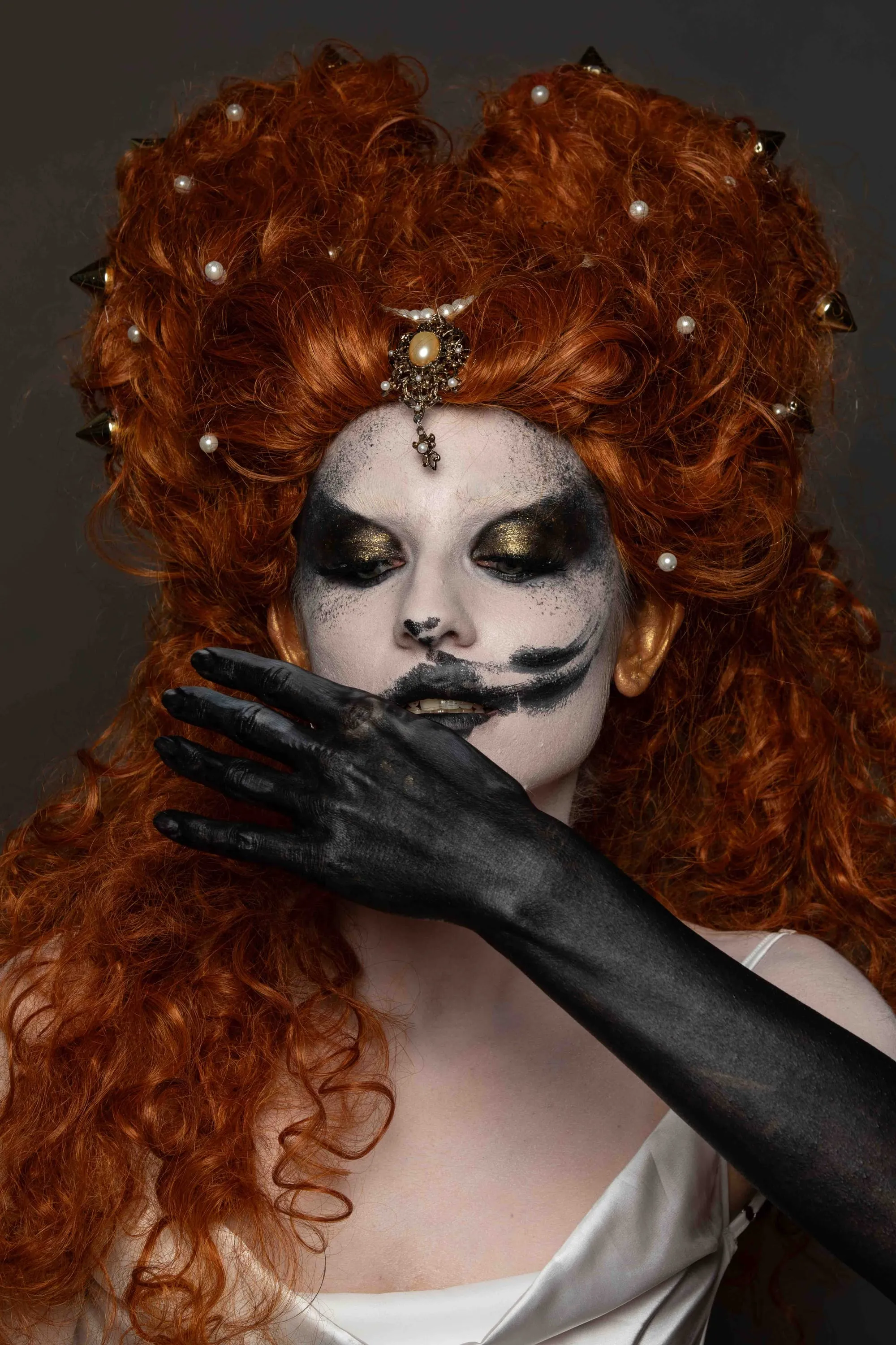 A red wig and theatrical make-up, a black hand covers the mouth
