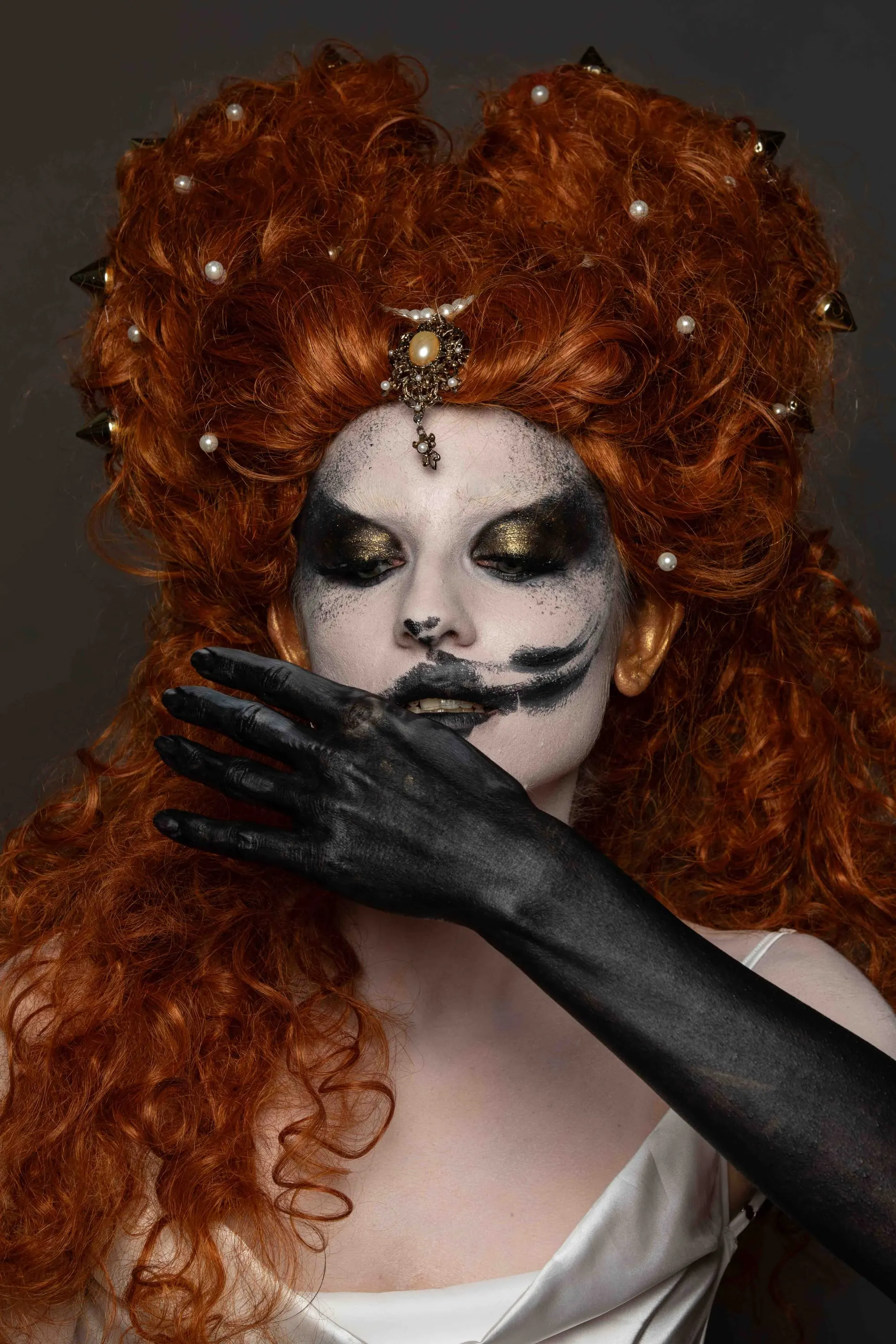 A red wig and theatrical make-up, a black hand covers the mouth