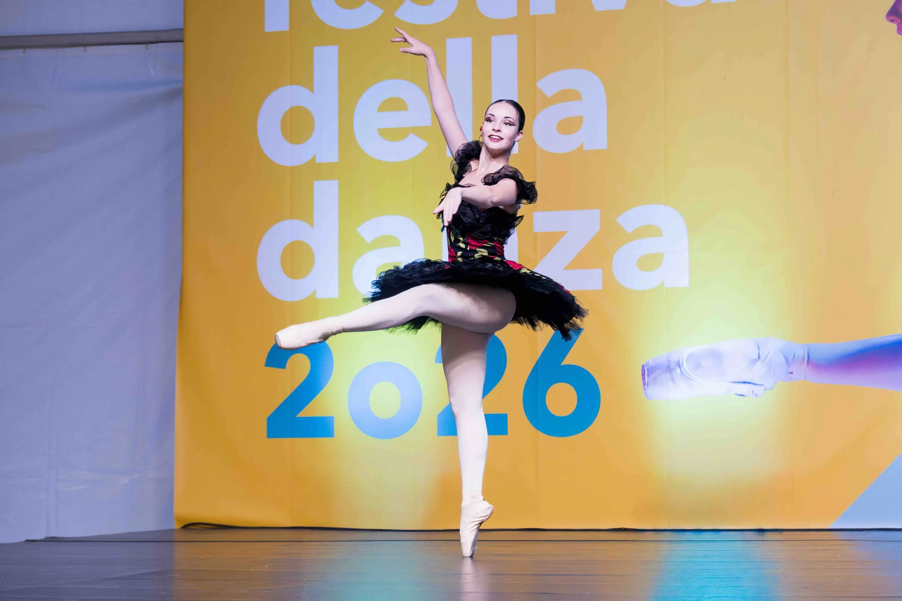 A ballerina in a black tutu in arabesque before the yellow festival backdrop