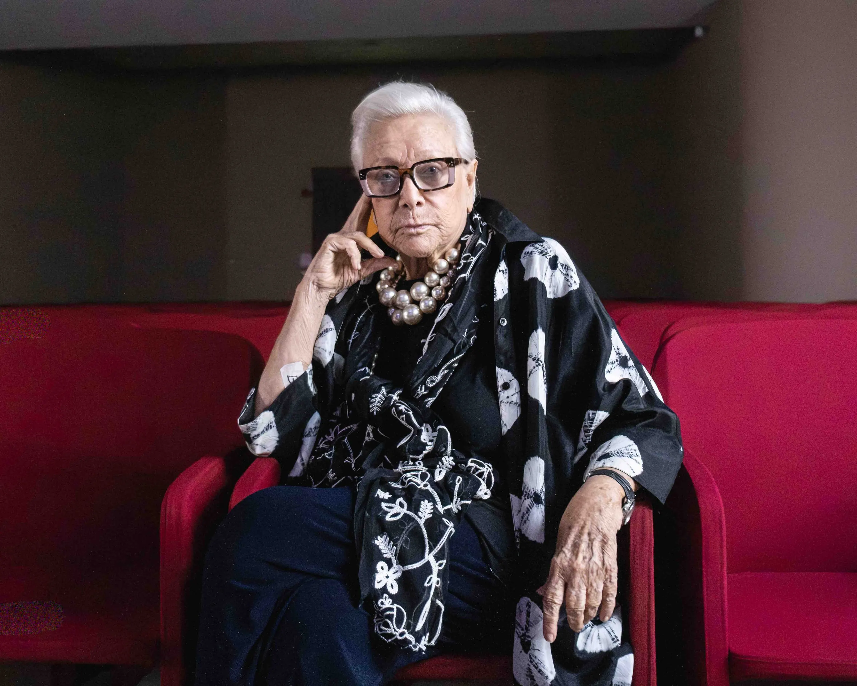 A white-haired woman in a floral shawl seated in a red armchair