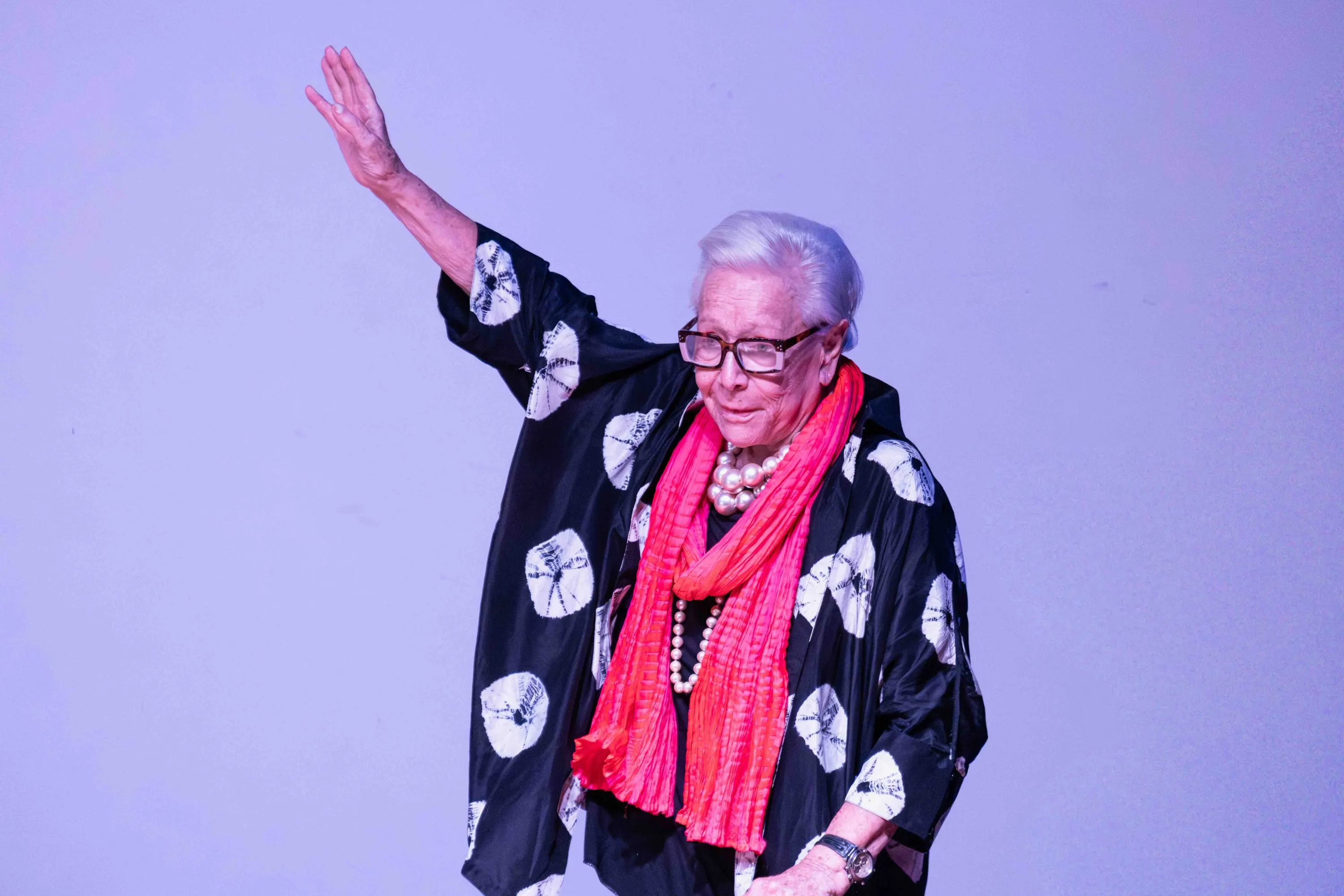 The white-haired woman waves on stage with her arm raised
