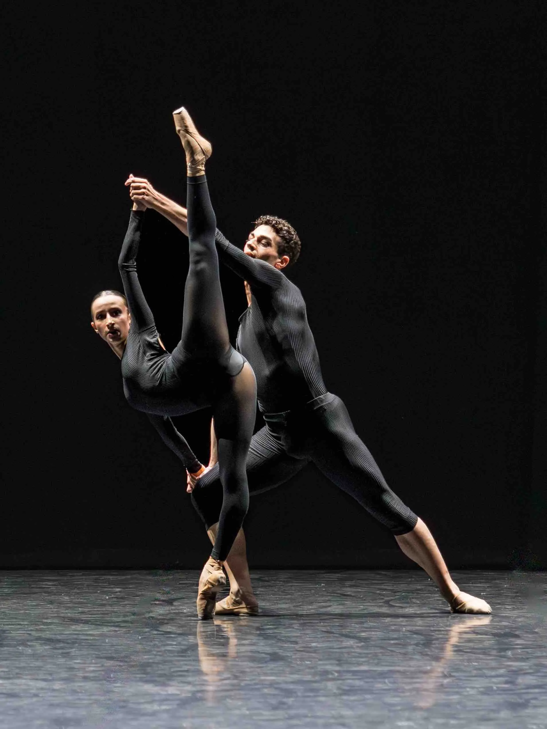 A male dancer supports a ballerina as she raises one leg straight up