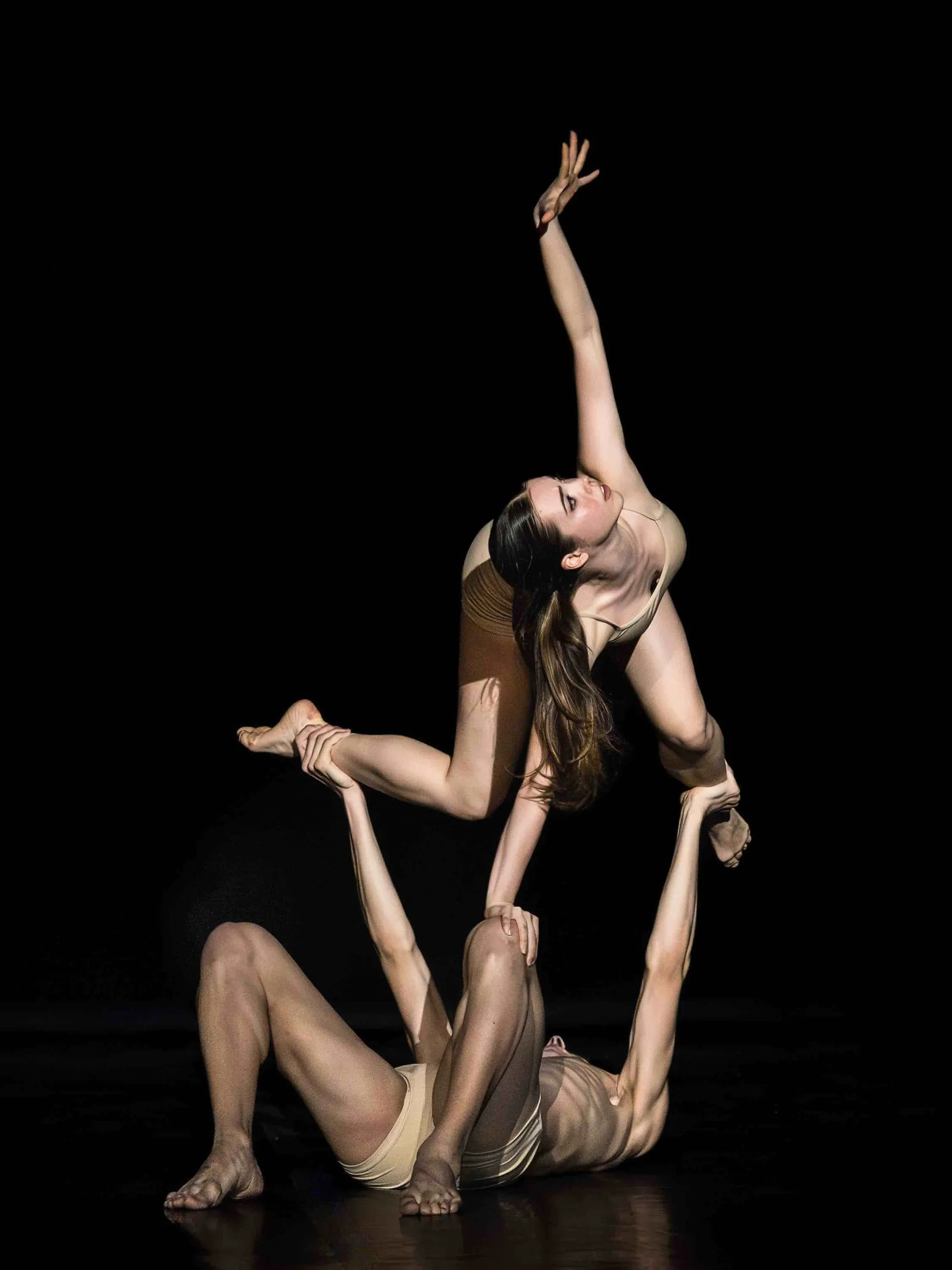 A dancer balances above her partner lying on the floor, black background