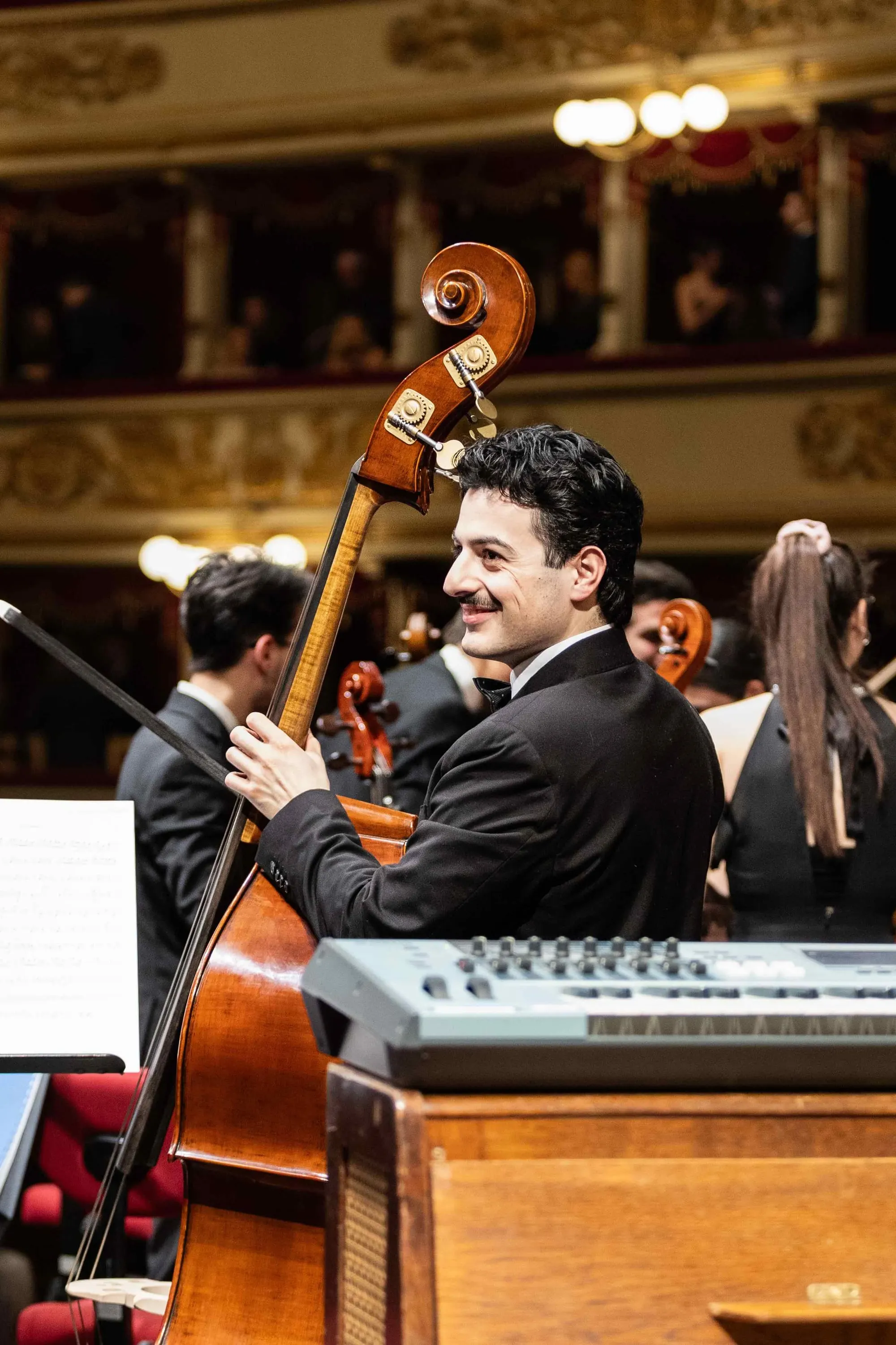 A double bass player smiles among the musicians, La Scala's boxes behind